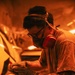 A tattooed male worker is using a power sander on what appears to be an automotive body panel in a workshop environment. He is wearing appropriate personal protective equipment including a dual-cartridge respirator mask, safety glasses, and work gloves. The scene is bathed in warm orange ambient lighting, likely from heat lamps used in auto body work.