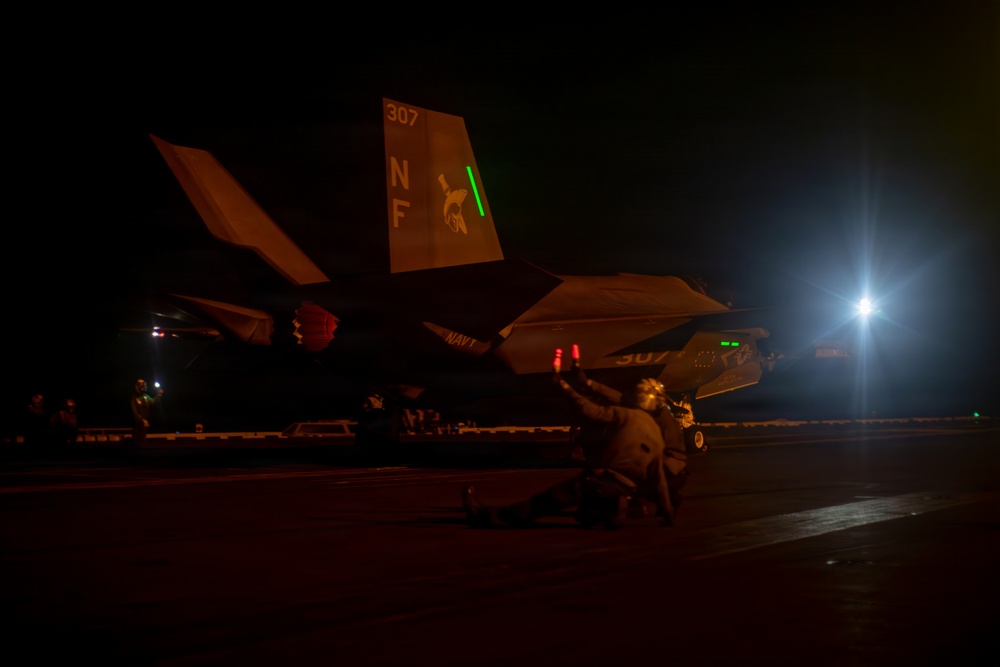 A U.S. Navy F-35C Lightning II stealth fighter jet (Bureau Number 307) is photographed on what appears to be an aircraft carrier flight deck during nighttime operations. The tail markings show 'NF' squadron codes and the number '307', with a squadron insignia visible on the tail fin. Ground crew personnel are visible working around the aircraft under low-light conditions.
