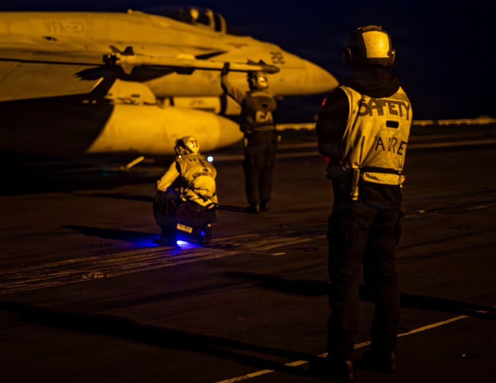 A nighttime scene on an aircraft carrier flight deck showing deck crew personnel conducting flight operations. The image is illuminated by warm amber/orange lighting, revealing at least three crew members near a military jet aircraft. A safety officer wearing a reflective vest marked 'SAFETY' and 'AIRE' is prominently visible in the foreground.