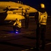 A nighttime scene on an aircraft carrier flight deck showing deck crew personnel conducting flight operations. The image is illuminated by warm amber/orange lighting, revealing at least three crew members near a military jet aircraft. A safety officer wearing a reflective vest marked 'SAFETY' and 'AIRE' is prominently visible in the foreground.
