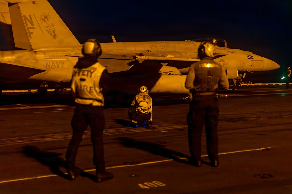 A nighttime flight deck operation aboard a U.S. Navy aircraft carrier, illuminated by warm artificial lighting. Three naval personnel are observing or conducting pre-flight/post-flight checks on an F/A-18 Hornet assigned to VFA-27, bureau number 212. The scene depicts standard carrier deck crew procedures during low-light operations.
