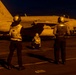 A nighttime flight deck operation aboard a U.S. Navy aircraft carrier, illuminated by warm artificial lighting. Three naval personnel are observing or conducting pre-flight/post-flight checks on an F/A-18 Hornet assigned to VFA-27, bureau number 212. The scene depicts standard carrier deck crew procedures during low-light operations.