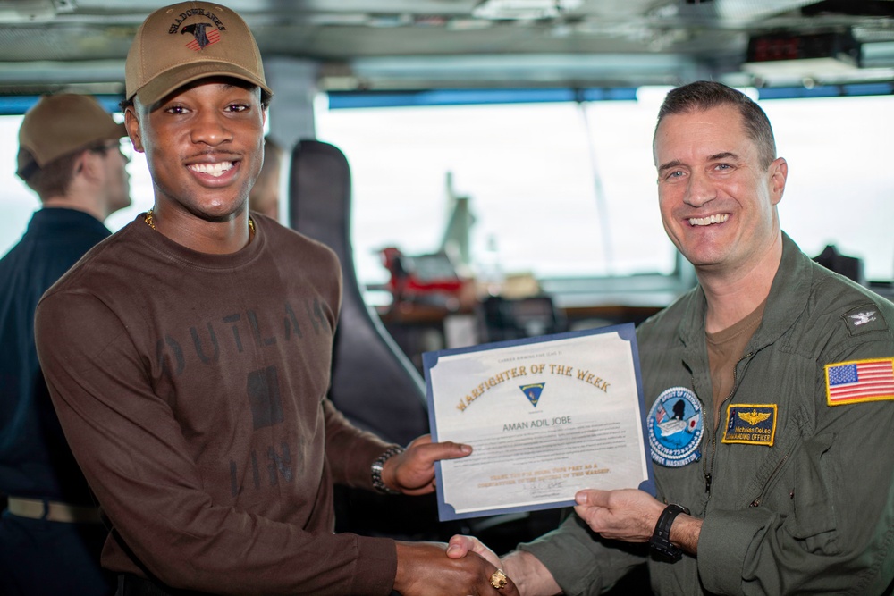 Two individuals are posing together while holding a 'Warfighter of the Week' certificate awarded to 'Aman Adil Jobe'. The man on the right is wearing a military flight suit with a U.S. flag patch and a name tag reading 'Commanding Officer', while the recipient on the left wears civilian clothing and a cap labeled 'Shadowhawks'. The setting appears to be aboard a naval vessel, likely on the bridge or command deck.