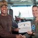 Two individuals are posing together while holding a 'Warfighter of the Week' certificate awarded to 'Aman Adil Jobe'. The man on the right is wearing a military flight suit with a U.S. flag patch and a name tag reading 'Commanding Officer', while the recipient on the left wears civilian clothing and a cap labeled 'Shadowhawks'. The setting appears to be aboard a naval vessel, likely on the bridge or command deck.