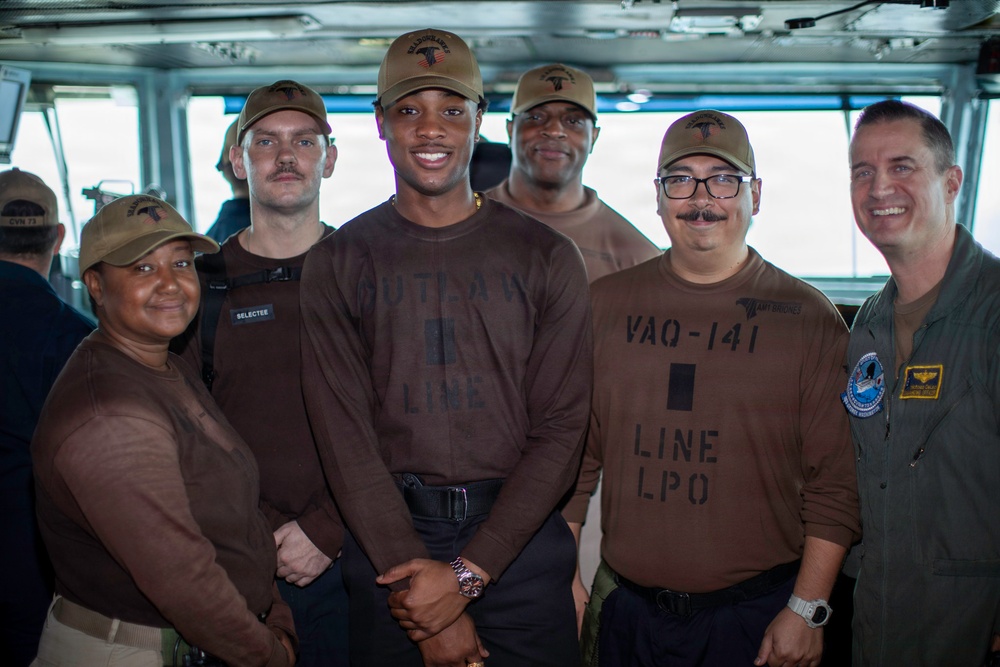 A group of six individuals posing for a photo aboard what appears to be a U.S. Navy vessel, likely on the bridge or an interior command area. Several individuals are wearing brown military-style shirts with unit markings, including 'VAQ-141 LINE LPO' and 'OUTLAW LINE', along with matching tan caps. One individual on the right is wearing a flight suit with squadron patches.