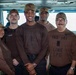 A group of six individuals posing for a photo aboard what appears to be a U.S. Navy vessel, likely on the bridge or an interior command area. Several individuals are wearing brown military-style shirts with unit markings, including 'VAQ-141 LINE LPO' and 'OUTLAW LINE', along with matching tan caps. One individual on the right is wearing a flight suit with squadron patches.