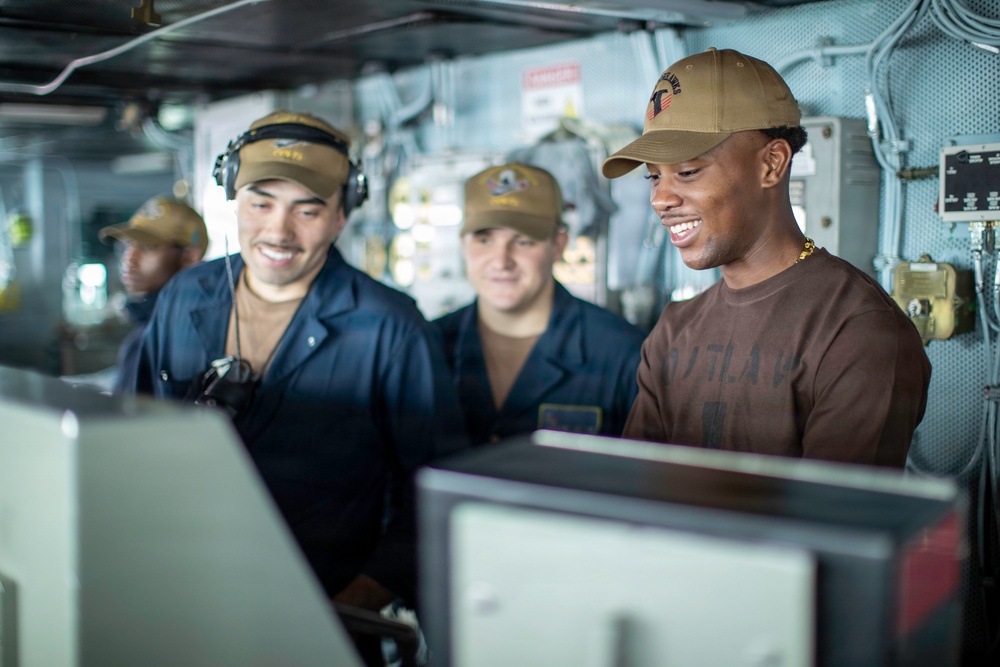 The image shows a group of individuals aboard what appears to be a military naval vessel, gathered around equipment consoles in a ship's interior compartment. At least four people are visible, with three in the foreground appearing engaged and smiling at a display or console. The setting is consistent with a ship's bridge or operations room, featuring visible wiring, panels, and naval equipment.