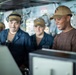 The image shows a group of individuals aboard what appears to be a military naval vessel, gathered around equipment consoles in a ship's interior compartment. At least four people are visible, with three in the foreground appearing engaged and smiling at a display or console. The setting is consistent with a ship's bridge or operations room, featuring visible wiring, panels, and naval equipment.