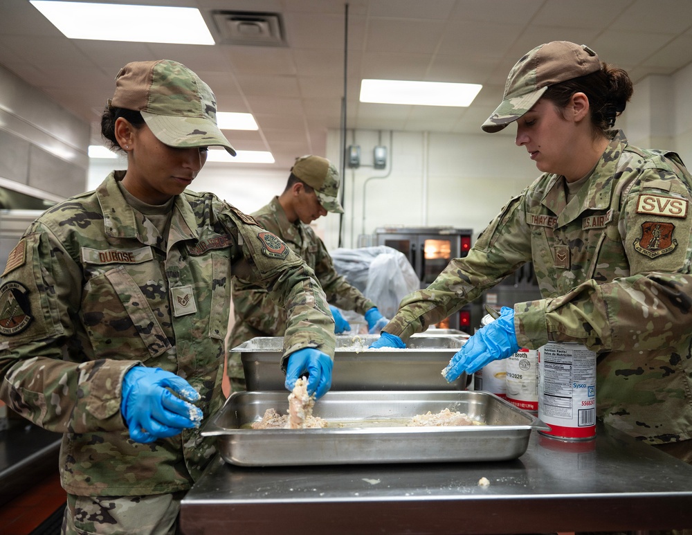 Three U.S. Air Force personnel in OCP (Operational Camouflage Pattern) uniforms are preparing food in a military kitchen facility. The airmen, identifiable by name tapes 'DUBOSE' and 'THAYER' and an 'SVS' (Services) patch, are handling what appears to be raw meat in stainless steel pans while wearing blue nitrile gloves. The scene depicts a food service operation consistent with a military dining facility (DFAC) environment.