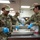 Three U.S. Air Force personnel in OCP (Operational Camouflage Pattern) uniforms are preparing food in a military kitchen facility. The airmen, identifiable by name tapes 'DUBOSE' and 'THAYER' and an 'SVS' (Services) patch, are handling what appears to be raw meat in stainless steel pans while wearing blue nitrile gloves. The scene depicts a food service operation consistent with a military dining facility (DFAC) environment.