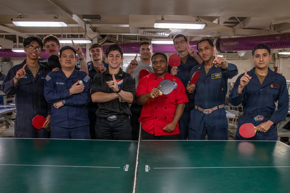 A group of approximately eleven individuals, most wearing U.S. Navy blue coverall uniforms, pose together around a green ping-pong table aboard what appears to be a naval vessel. Several members are holding table tennis paddles and raising index fingers in a 'number one' gesture, suggesting a tournament or competition victory celebration. One individual stands out wearing a red chef's jacket, and another wears a black shirt.