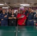 A group of approximately eleven individuals, most wearing U.S. Navy blue coverall uniforms, pose together around a green ping-pong table aboard what appears to be a naval vessel. Several members are holding table tennis paddles and raising index fingers in a 'number one' gesture, suggesting a tournament or competition victory celebration. One individual stands out wearing a red chef's jacket, and another wears a black shirt.