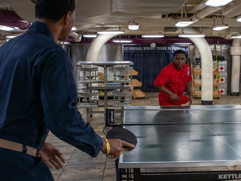 Two individuals are playing table tennis (ping pong) in what appears to be a below-deck recreational area of a naval or military vessel. One player wears a navy blue uniform and holds a paddle, while the other in a red shirt is positioned on the opposite side of a Kettler brand table. The space features industrial piping, stacked metal furniture, Red Cross signage, and a motivational banner on the wall.