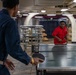 Two individuals are playing table tennis (ping pong) in what appears to be a below-deck recreational area of a naval or military vessel. One player wears a navy blue uniform and holds a paddle, while the other in a red shirt is positioned on the opposite side of a Kettler brand table. The space features industrial piping, stacked metal furniture, Red Cross signage, and a motivational banner on the wall.