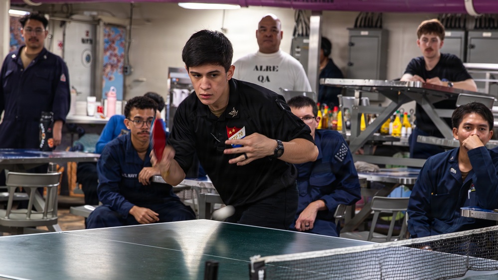 A young man in a black uniform is actively playing table tennis (ping pong) aboard what appears to be a U.S. Navy vessel, likely in a mess hall or recreation area. He is mid-swing with a red paddle while several other personnel in navy blue coveralls watch from the background. The setting suggests a morale, welfare, and recreation (MWR) activity for military personnel.