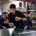 A young man in a black uniform is actively playing table tennis (ping pong) aboard what appears to be a U.S. Navy vessel, likely in a mess hall or recreation area. He is mid-swing with a red paddle while several other personnel in navy blue coveralls watch from the background. The setting suggests a morale, welfare, and recreation (MWR) activity for military personnel.