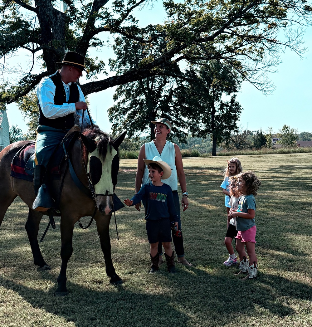 A man dressed in period costume (appearing to be Civil War-era attire) sits atop a dark horse with a white blaze, interacting with a woman and three children on a grassy open field. The children, one wearing a cowboy hat and boots, appear engaged and curious about the horse. The setting suggests a historical reenactment event or living history demonstration at an outdoor venue.