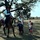 A man dressed in period costume (appearing to be Civil War-era attire) sits atop a dark horse with a white blaze, interacting with a woman and three children on a grassy open field. The children, one wearing a cowboy hat and boots, appear engaged and curious about the horse. The setting suggests a historical reenactment event or living history demonstration at an outdoor venue.