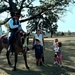 A man dressed in period costume (appearing to be Civil War-era attire) sits atop a dark horse with a white blaze, interacting with a woman and three children on a grassy open field. The children, one wearing a cowboy hat and boots, appear engaged and curious about the horse. The setting suggests a historical reenactment event or living history demonstration at an outdoor venue.