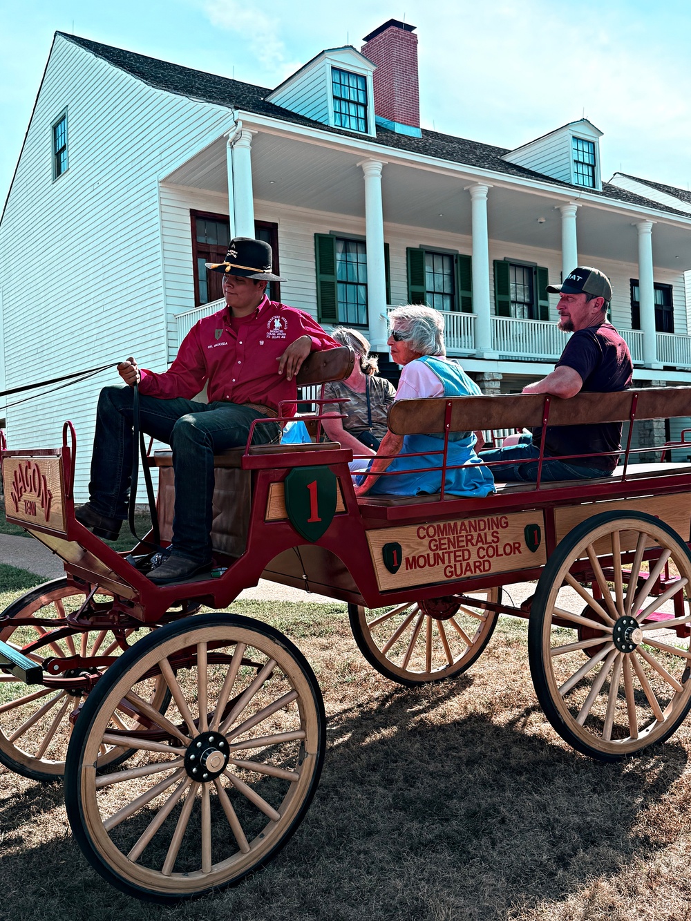 A red wooden wagon labeled 'Commanding Generals Mounted Color Guard' with the number '1' and a green shield insignia is carrying several passengers near a historic white colonial-style building. The driver, wearing a red shirt and black hat, sits at the front holding reins. This appears to be a historical tour or ceremonial event at a military installation or heritage site.