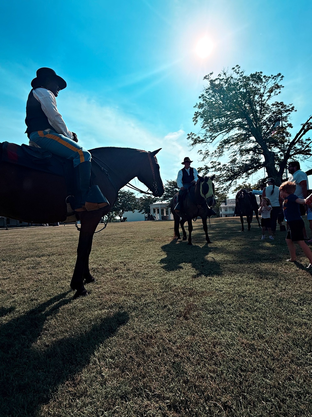 An outdoor historical reenactment or equestrian event taking place on a grassy field, likely at a historic site given the white antebellum-style building visible in the background. Multiple riders on horseback are present, wearing period-style clothing including wide-brimmed hats and cavalry-style trousers with stripe detailing. Spectators, including children, are gathered nearby interacting with the riders and horses.
