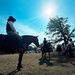An outdoor historical reenactment or equestrian event taking place on a grassy field, likely at a historic site given the white antebellum-style building visible in the background. Multiple riders on horseback are present, wearing period-style clothing including wide-brimmed hats and cavalry-style trousers with stripe detailing. Spectators, including children, are gathered nearby interacting with the riders and horses.