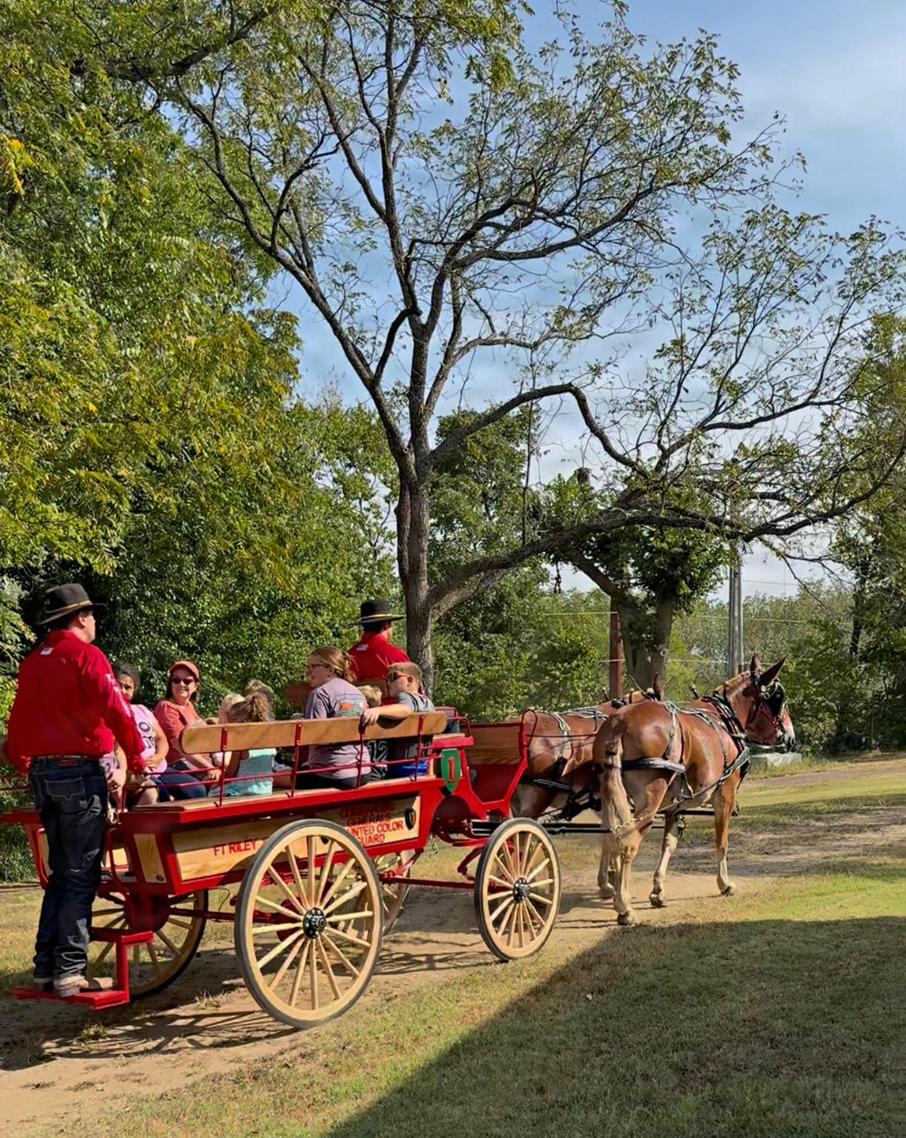 A bright red horse-drawn wagon associated with 'FT Riley Mounted Color Guard' carries a group of passengers along a dirt path surrounded by trees. Two individuals wearing red shirts and black cowboy hats appear to be guides or handlers accompanying the wagon. The scene appears to be a recreational or ceremonial wagon ride taking place outdoors in a park-like setting.