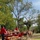 A bright red horse-drawn wagon associated with 'FT Riley Mounted Color Guard' carries a group of passengers along a dirt path surrounded by trees. Two individuals wearing red shirts and black cowboy hats appear to be guides or handlers accompanying the wagon. The scene appears to be a recreational or ceremonial wagon ride taking place outdoors in a park-like setting.