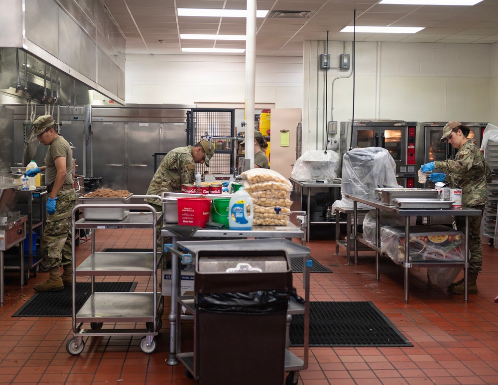 Military personnel in camouflage uniforms and blue food-service gloves are working in a large institutional kitchen, preparing food at stainless steel prep tables. The kitchen is well-equipped with commercial ovens, refrigeration units, and ventilation hoods. Multiple soldiers are engaged in food preparation tasks simultaneously, suggesting a large-scale meal service operation.
