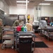 Military personnel in camouflage uniforms and blue food-service gloves are working in a large institutional kitchen, preparing food at stainless steel prep tables. The kitchen is well-equipped with commercial ovens, refrigeration units, and ventilation hoods. Multiple soldiers are engaged in food preparation tasks simultaneously, suggesting a large-scale meal service operation.