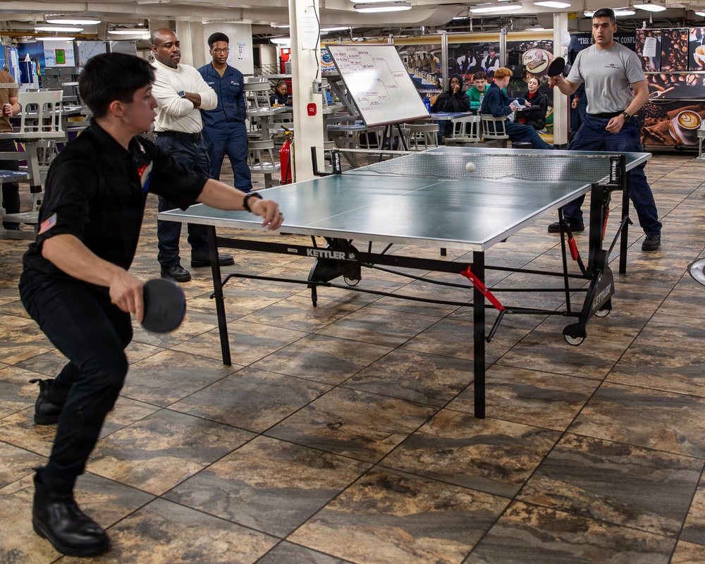 Two individuals are engaged in a competitive table tennis match on a Kettler brand ping-pong table inside what appears to be a military vessel's mess hall or recreation area. The player in the foreground wears a black uniform with a US flag patch and is in a dynamic hitting stance, while the opponent in a gray shirt stands on the opposite side. Several spectators, some in military uniforms, observe the match in the background.
