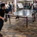Two individuals are engaged in a competitive table tennis match on a Kettler brand ping-pong table inside what appears to be a military vessel's mess hall or recreation area. The player in the foreground wears a black uniform with a US flag patch and is in a dynamic hitting stance, while the opponent in a gray shirt stands on the opposite side. Several spectators, some in military uniforms, observe the match in the background.