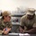 Two military personnel in OCP (Operational Camouflage Pattern) uniforms are engaged in a discussion at a stainless steel counter in what appears to be a military dining facility or kitchen. The female service member on the left is speaking and gesturing while holding a small blue object, while the male service member on the right is writing on a document with a pen. The setting suggests an administrative or operational briefing taking place in a food service environment.