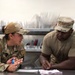 Two military personnel in OCP (Operational Camouflage Pattern) uniforms are engaged in a discussion at a stainless steel counter in what appears to be a military dining facility or kitchen. The female service member on the left is speaking and gesturing while holding a small blue object, while the male service member on the right is writing on a document with a pen. The setting suggests an administrative or operational briefing taking place in a food service environment.