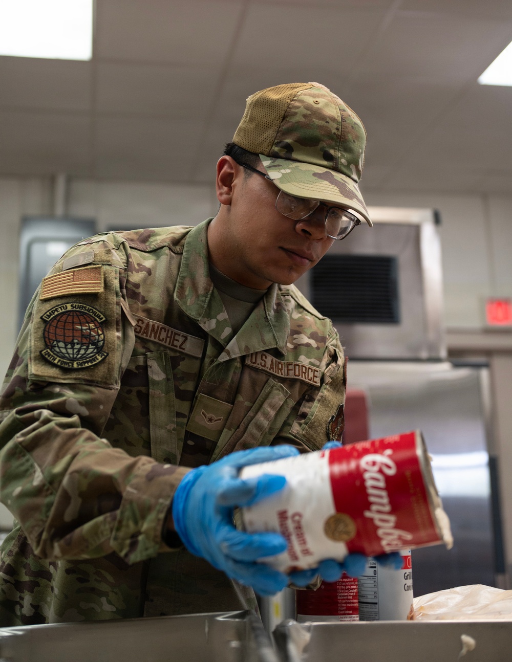 A U.S. Air Force service member named Sanchez, wearing OCP (Operational Camouflage Pattern) uniform, is working in a military kitchen facility. The airman is handling a Campbell's soup can while wearing blue nitrile gloves and safety glasses, suggesting food preparation duties. The setting appears to be a military dining facility (DFAC) or mess hall.