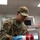 A U.S. Air Force service member named Sanchez, wearing OCP (Operational Camouflage Pattern) uniform, is working in a military kitchen facility. The airman is handling a Campbell's soup can while wearing blue nitrile gloves and safety glasses, suggesting food preparation duties. The setting appears to be a military dining facility (DFAC) or mess hall.