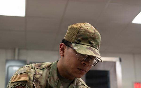 A U.S. Air Force service member named Sanchez, wearing OCP (Operational Camouflage Pattern) uniform, is working in a military kitchen facility. The airman is handling a Campbell's soup can while wearing blue nitrile gloves and safety glasses, suggesting food preparation duties. The setting appears to be a military dining facility (DFAC) or mess hall.