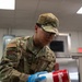 A U.S. Air Force service member named Sanchez, wearing OCP (Operational Camouflage Pattern) uniform, is working in a military kitchen facility. The airman is handling a Campbell's soup can while wearing blue nitrile gloves and safety glasses, suggesting food preparation duties. The setting appears to be a military dining facility (DFAC) or mess hall.