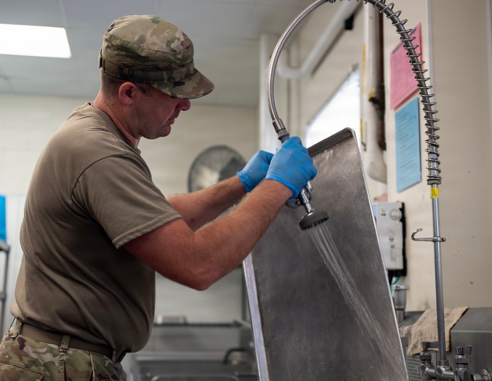 A military service member in camouflage uniform and blue latex gloves is rinsing or cleaning a large stainless steel tray using a commercial spray hose in what appears to be a military dining facility kitchen. The individual is wearing an OCP (Operational Camouflage Pattern) uniform with a matching cap and an olive drab t-shirt. The setting is an institutional kitchen environment with visible equipment and posted notices on the wall.