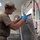 A military service member in camouflage uniform and blue latex gloves is rinsing or cleaning a large stainless steel tray using a commercial spray hose in what appears to be a military dining facility kitchen. The individual is wearing an OCP (Operational Camouflage Pattern) uniform with a matching cap and an olive drab t-shirt. The setting is an institutional kitchen environment with visible equipment and posted notices on the wall.