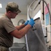 A military service member in camouflage uniform and blue latex gloves is rinsing or cleaning a large stainless steel tray using a commercial spray hose in what appears to be a military dining facility kitchen. The individual is wearing an OCP (Operational Camouflage Pattern) uniform with a matching cap and an olive drab t-shirt. The setting is an institutional kitchen environment with visible equipment and posted notices on the wall.