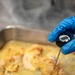 A food service worker wearing a blue nitrile glove is using an AVATEMP digital instant-read thermometer to check the internal temperature of cooked chicken in a stainless steel baking pan. The thermometer display appears to read approximately 165°F, which is the USDA-recommended safe internal temperature for poultry. This appears to be a food safety compliance check in a commercial or institutional kitchen setting.