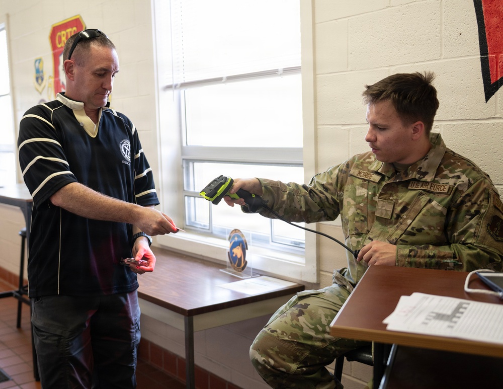 A U.S. Air Force servicemember named McBride is seated at a desk scanning what appears to be an ID card or badge belonging to a civilian man in a dark polo shirt. The interaction appears to be an access control or check-in procedure taking place inside a military facility. Military unit patches and insignia are visible on the wall in the background.