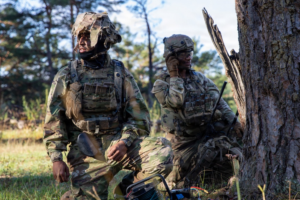 Two soldiers in MultiCam/OCP camouflage uniforms are positioned near a large tree in a forested outdoor environment. The soldier on the left wears a ghillie-style camouflaged helmet cover and tactical vest, while the soldier on the right appears to be communicating via radio or earpiece. A piece of equipment, possibly a radio or generator, is visible on the ground between them.