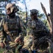 Two soldiers in MultiCam/OCP camouflage uniforms are positioned near a large tree in a forested outdoor environment. The soldier on the left wears a ghillie-style camouflaged helmet cover and tactical vest, while the soldier on the right appears to be communicating via radio or earpiece. A piece of equipment, possibly a radio or generator, is visible on the ground between them.