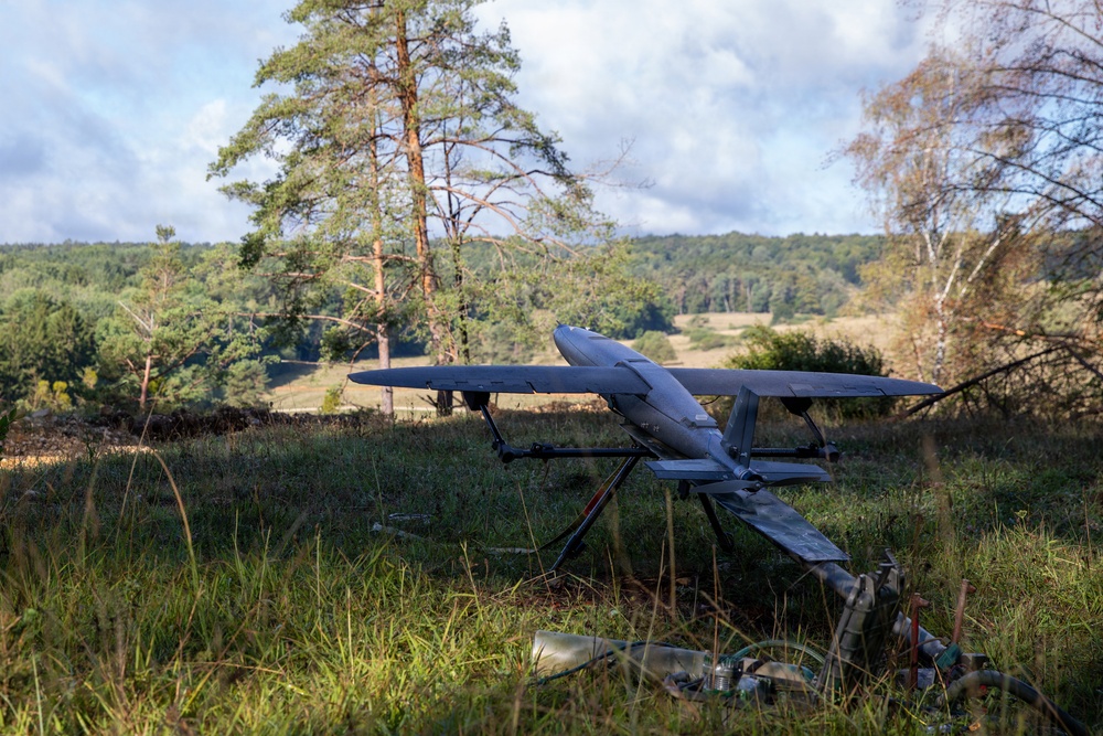 A fixed-wing unmanned aerial vehicle (UAV) is positioned on a launch or ground support frame in an open grassy field surrounded by trees. The drone appears to be a military or reconnaissance-type aircraft with a gray finish, mounted on a metal stand. Various equipment and supplies are visible on the ground nearby, suggesting a field deployment or pre-launch setup.
