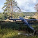 A fixed-wing unmanned aerial vehicle (UAV) is positioned on a launch or ground support frame in an open grassy field surrounded by trees. The drone appears to be a military or reconnaissance-type aircraft with a gray finish, mounted on a metal stand. Various equipment and supplies are visible on the ground nearby, suggesting a field deployment or pre-launch setup.