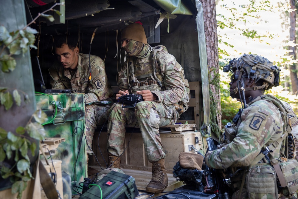 Three U.S. Army soldiers in Operational Camouflage Pattern (OCP) uniforms are operating from what appears to be a military vehicle or command post in a wooded area. Two soldiers are seated inside the vehicle working with electronic equipment and a handheld controller, while a third soldier in full combat gear stands guard nearby. The setting suggests a field exercise or tactical operation.