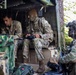 Three U.S. Army soldiers in Operational Camouflage Pattern (OCP) uniforms are operating from what appears to be a military vehicle or command post in a wooded area. Two soldiers are seated inside the vehicle working with electronic equipment and a handheld controller, while a third soldier in full combat gear stands guard nearby. The setting suggests a field exercise or tactical operation.