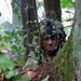 A soldier in full camouflage gear is concealed among trees and foliage in a woodland environment. The individual is wearing a helmet adorned with natural vegetation and equipped with night-vision device mounts. The image depicts a tactical concealment or reconnaissance posture in a dense forest setting.